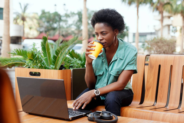 Μια όμορφη Αφρικανή πίνει καφέ σε ένα καφέ και εργάζεται σε ένα λάπτοπ, ακουστικά και ένα τηλέφωνο βρίσκονται δίπλα της. Το κορίτσι δουλεύει στον υπολογιστή κατά τη διάρκεια ενός διαλείμματος καφέ. - Φωτογραφία, εικόνα