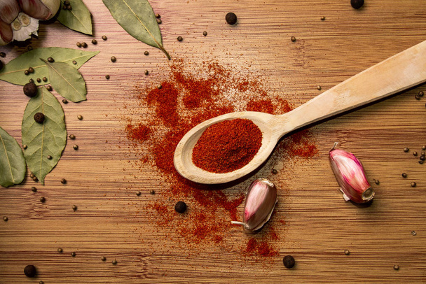 Ground chili in a char spoon on a wooden background. View from above. Garlic and bay leaf on the background. Seasonings for meat and spicy dishes.. - Photo, image
