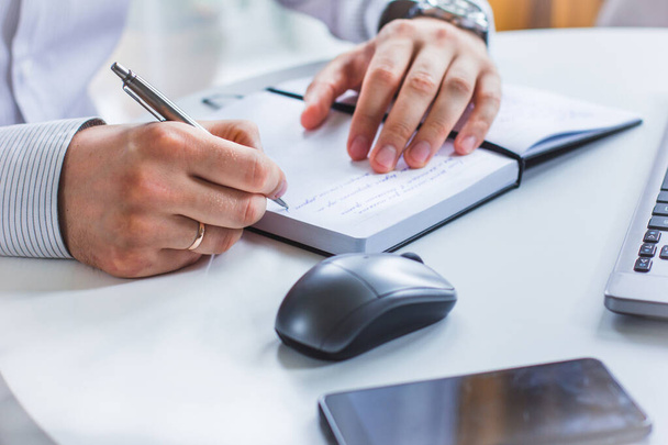 An image of the hands of a man sitting at a table and writing in a Notepad with a ballpoint pen. Next to it is a laptop, a computer mouse, and a cell phone - 写真・画像