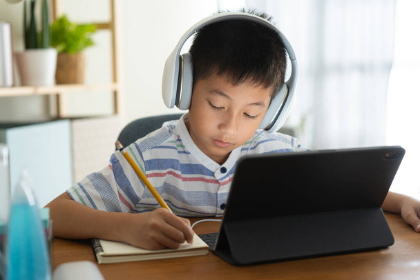 Азіатський хлопчик навчається онлайн через Інтернет з учителем на планшеті цифрового з навушником, Азіатська дитина вчиться, сидячи в вітальні вдома. Концепція онлайн-навчання вдома.  - Фото, зображення