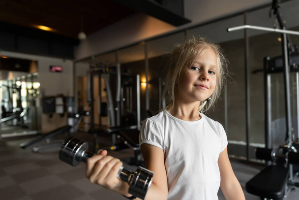 Little cute adorable caucasian blond girl doing finess exercise lifting dumbbell in sport gym club indoors. Happy sporty tired child. Children healthcare workout and healthy lifestyle concept - Photo, Image