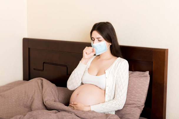 Беременная женщина в защитной маске. COVID-19 Coronavirus сидит на диване. Предотвратить заражение плода. - Фото, изображение