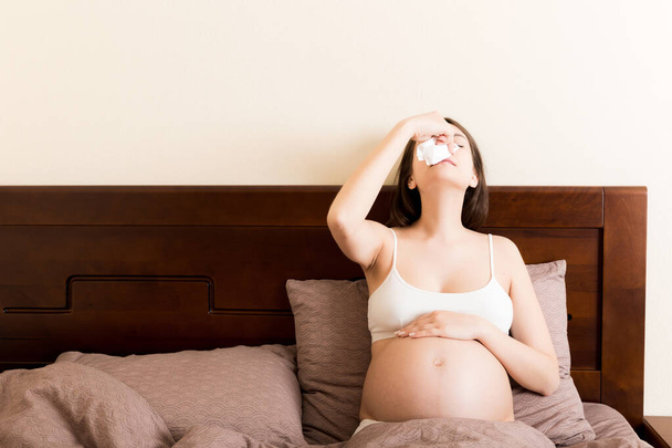 Porträt einer jungen unglücklichen Schwangeren, die sich auf Kissen auf dem Bett lehnt und ihre Nase in Gewebe pustet. Künftige Mama erkältet und niest. - Foto, Bild