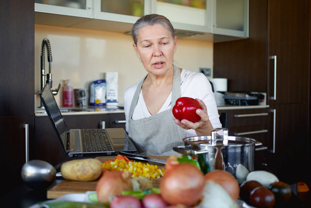 Зрелая женщина на кухне готовит еду и пользуется ноутбуком. - Фото, изображение