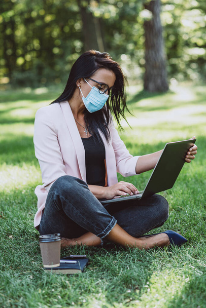 Молодая женщина в защитной маске использует ноутбук в общественном парке во время работы на открытом воздухе. Дистанционное обучение и телеработа во время пандемии. Мягкая селективная фокусировка. - Фото, изображение