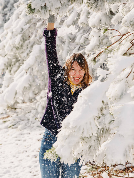 Portrait of young woman among snowy trees in winter forest. Smiling woman playing with snow in sunny cold day - Foto, Imagen