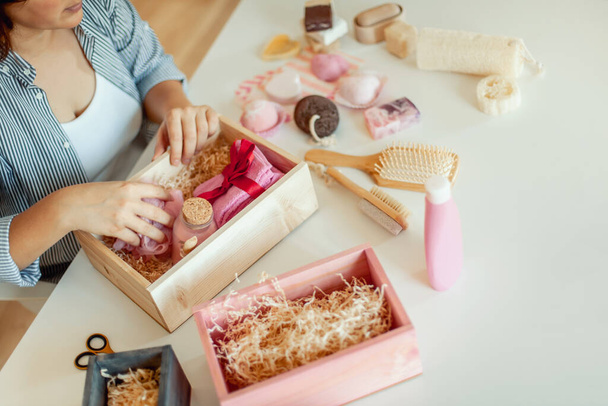Young woman placing handmade organic soaps, bath salts, sponge with brush and assortment of bath bombs into bridesmaid gift box set. Concept of beauty treatment gift boxes for women to make her day. - 写真・画像