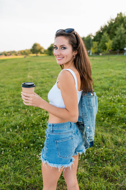 Happy woman in summer in park holds cup with coffee tea hands, meadows, enjoys rest. Emotions of tenderness, pleasure joy, breakfast in nature. White tank top tanned leather, denim shorts and jacket. - Fotografie, Obrázek