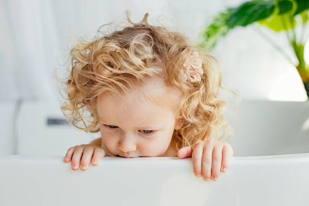 Una pequeña chica de pelo rizado con cabello rubio está sentada en el baño. Bromas y juegos infantiles. - Foto, Imagen