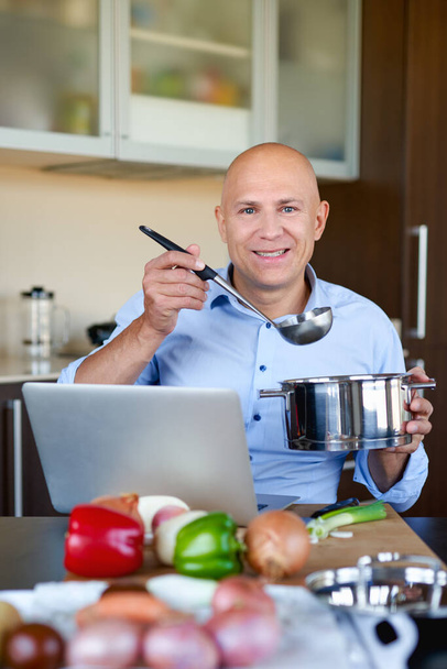 Человек на кухне ищет рецепты на своем ноутбуке. Кулинария в доме, онлайн - Фото, изображение