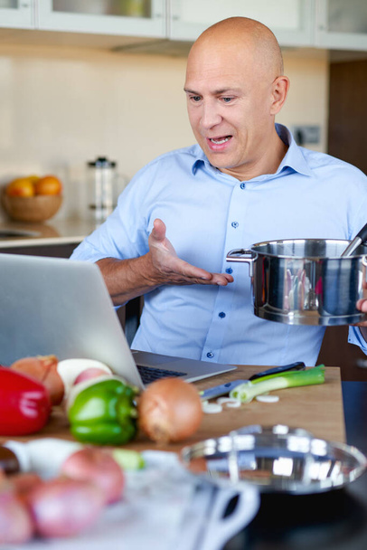 Yetişkin vahşi adam mutfakta yemek hazırlıyor ve dizüstü bilgisayara bakıyor.. - Fotoğraf, Görsel