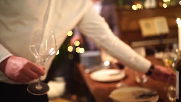 Χαμηλό φως. Ένας άντρας με λευκό πουκάμισο βάζει ποτήρια κρασιού στο τραπέζι. δείπνο υπό το φως κεριών - Πλάνα, βίντεο