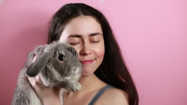 Портрет щасливої молодої жінки, що обіймає і цілує декоративного сірого лопатевого кролика. Рожевий фон. Концепція захисту домашніх тварин
. - Кадри, відео