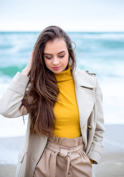 elegant brunette female with blue eyes in the coat on the winter windy beach, turquoise sea waves at the background - 写真・画像