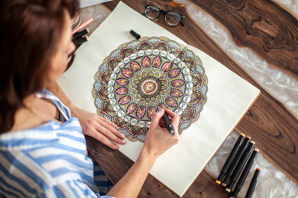 Pretty young woman coloring the mandala with colored markers. An album with a circular pattern lies on the table. Hobby and relaxation concept. - Photo, Image