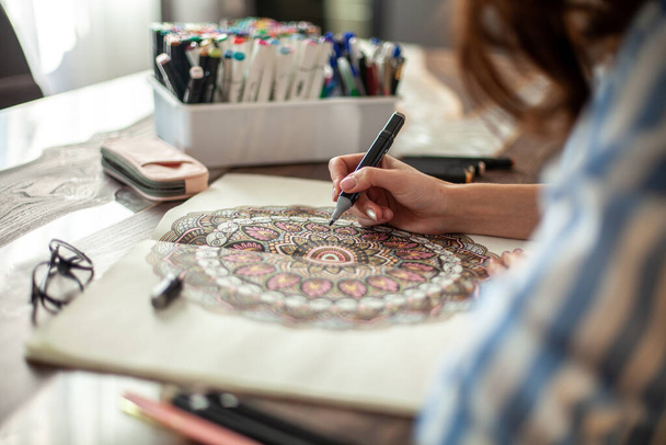 Beautiful well-groomed young woman draws a circular mandala pattern in the album. Artistic markers and colored pencils. Hobby, therapy and home recreation concept. - Foto, Bild