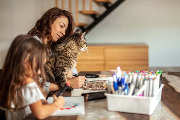 A young, slim and beautiful mother with a cute daughter and a Maine Coon cat is painting in the kitchen. Circular pattern mandala and zen drawing. Home entertainment, drawing and hobbies concept. Doodle drawing. Stylish kitchen. - Fotó, kép