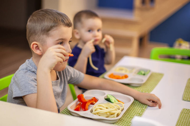 Two boys eat pasta, cucumber and sweet bell pepper. Home kitchen in the house. The older brother looks at the younger. Children are having dinner. - Fotoğraf, Görsel