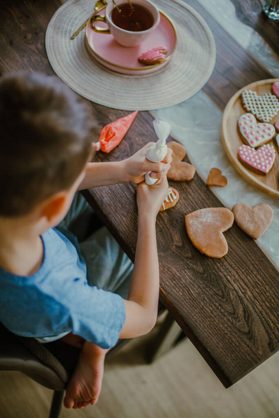 Little cute boy decorates gingerbread heart shaped with pink sugar icing. Preparation for Valentines day concept. Eats sweet cornet icing sugar. - Foto, Imagem