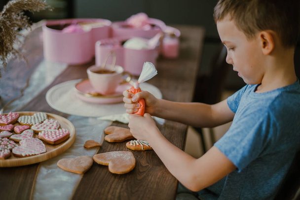 Little cute boy decorates gingerbread heart shaped with pink sugar icing. Preparation for Valentines day concept. Eats sweet cornet icing sugar. - Fotografie, Obrázek