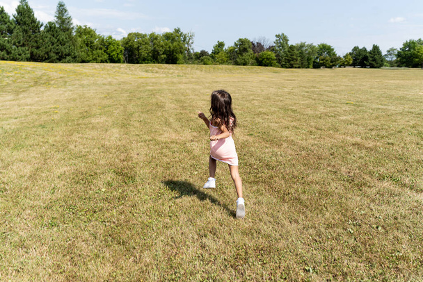 Νεαρό κορίτσι με ροζ φόρεμα και τρέχει σε ένα πάρκο που φαίνεται από πίσω - Φωτογραφία, εικόνα