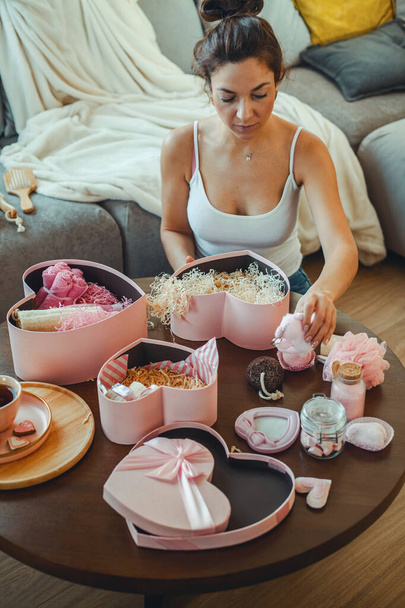 Young beautiful woman opens pink colored heart shaped gift boxes with homemade bath supplies, marshmallows and body care cosmetics products for woman on white marble background. Concept of Valentines day gifts. - Fotó, kép