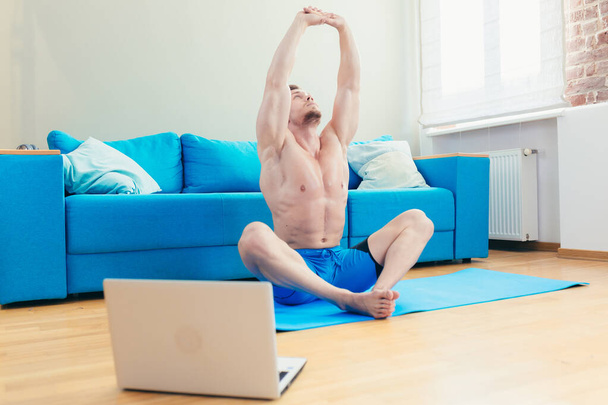 A muscular athletic man in shorts is energetically starting to do fitness exercises at home in his apartment with a modern interior. Classes online - Foto, Imagen