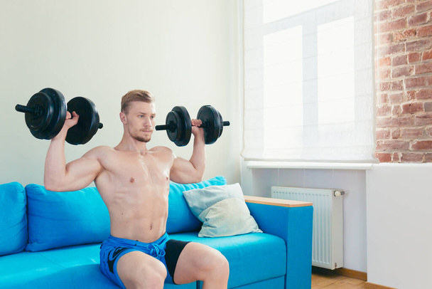 A muscular athletic man in shorts is energetically starting to do fitness exercises with dumbbells at home in his apartment with a modern interior. - Foto, Bild