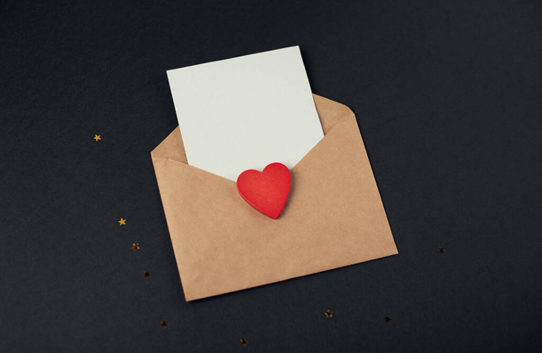 Sobre artesanal con una hoja de papel en blanco en el interior y corazón de madera roja en el fondo negro. Carta de amor romántica para el concepto de San Valentín. Espacio para texto. - Foto, imagen