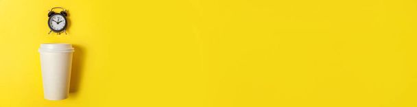 Simplemente diseño minimalista taza de café de papel y reloj despertador sobre fondo de color amarillo. Bebida para llevar y bebida para el desayuno. Buenos días, despierta concepto despierto. Banner plano vista superior copia espacio - Foto, imagen