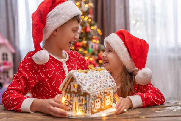 irmão e irmã em um chapéu de Papai Noel segurar uma casa de gengibre juntos. um quarto lindamente decorado com uma árvore de Natal, um menino e uma menina olham um para o outro e riem. Celebração do Natal - Foto, Imagem