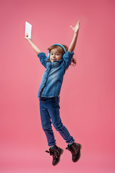Smiling little girl in headphones and with a tablet computer, jumping on a pink background. The child listens to music, plays and learns online. Electronic digital training. Dynamic image - Фото, зображення