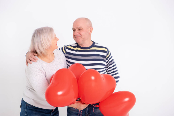 старша пара обіймається тримаючи червоні серцеві кульки
 - Фото, зображення