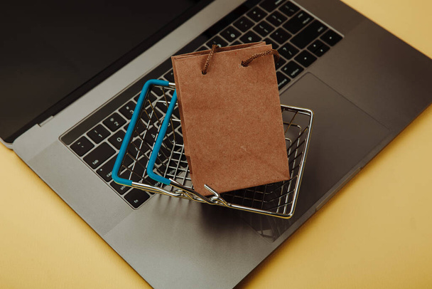 Konzept des Online-Shoppings. Papier Einkaufstasche und Einkaufswagen auf der Tastatur des Laptops isoliert auf gelb. Ansicht von oben - Foto, Bild