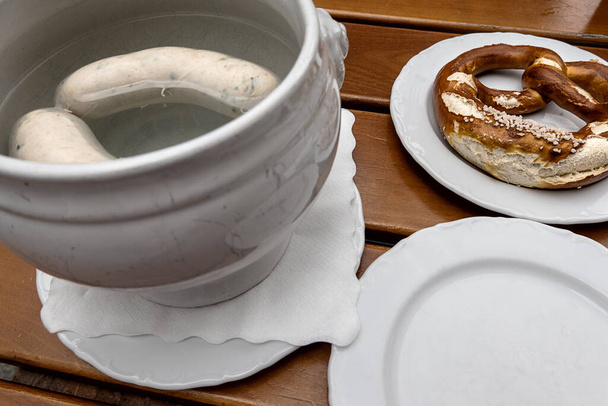 duas salsichas brancas bávaras em água quente, um pretzel em um prato e um prato vazio - Foto, Imagem