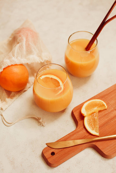Апельсиновый сок - апельсины в многоразовой торговой сумке со стеклянной кружкой и соломой из нержавеющей стали и деревянной доской для резки на белом столе - Фото, изображение