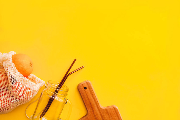 Апельсиновый сок плоский укладка - апельсины в многоразовой торговой сумке со стеклянной кружкой и соломинками из нержавеющей стали и деревянной резки доски на желтом фоне - Фото, изображение