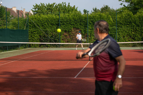 Ενεργοί ηλικιωμένοι που παίζουν τένις στο γήπεδο κατά τη διάρκεια της όμορφης καλοκαιρινής ημέρας, ένας ενεργός συνταξιούχος - Φωτογραφία, εικόνα