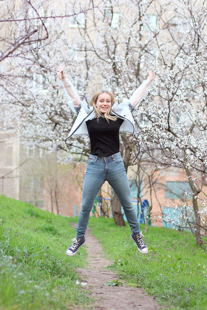 Una hermosa joven en pantalones vaqueros y zapatillas de deporte saltando a lo largo del camino contra el fondo de los árboles blancos florecientes - Foto, imagen