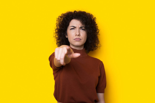 Сумна кучерява жінка вказує на камеру на жовтій стіні жестикулюючи нещастя
 - Фото, зображення