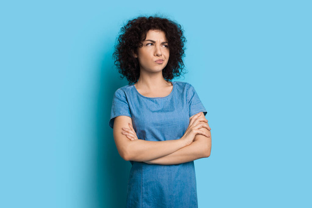 Curly haired woman is thinking a bout something gesturing it with her crossed hands and lips on a blue studio wall - Photo, Image