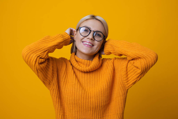 Monokróm fotó egy szőke nőről szemüvegben, aki megérinti a haját és mosolyog egy sárga stúdió falán. - Fotó, kép