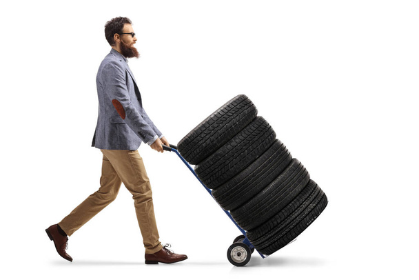 Full length profile shot of a bearded man pushing a hand truck with car tires isolated on white background - Zdjęcie, obraz