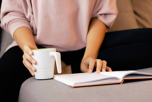 Αγνώριστη γυναίκα ανάγνωση βιβλίο, ενώ έχοντας φλιτζάνι τσάι και χαλάρωση στο σπίτι.  - Φωτογραφία, εικόνα