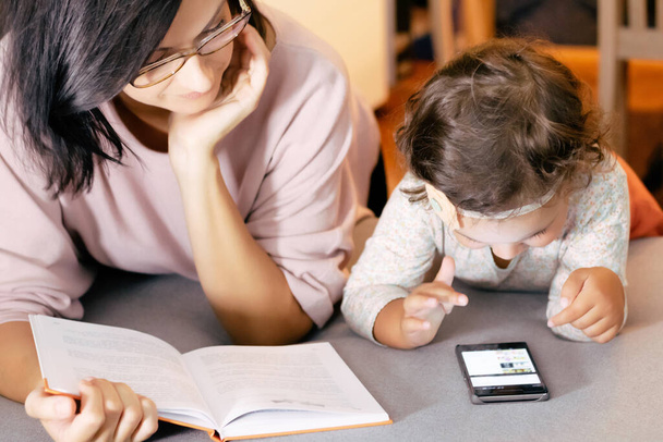 Маленькая девочка использует смартфон, пока ее мать читает книгу дома.  - Фото, изображение