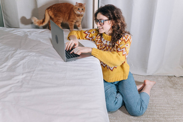 Stijlvolle vrouwelijke freelancer werkt thuis op een laptop zittend op de vloer met haar huisdier rode kat. - Foto, afbeelding