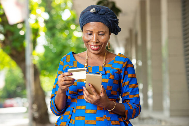 beautiful african businesswoman in traditional outfit standing outdoors holding mobile phone and credit card smiling. - Photo, Image