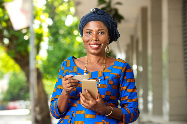 bella donna d'affari africana in abito tradizionale in piedi all'aperto con telefono cellulare e carta di credito sorridente. - Foto, immagini