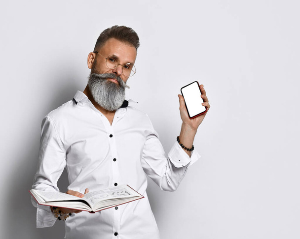 stylish brutal man with a gray beard holds a book or diary in his hands and shows the screen of his mobile phone - Fotoğraf, Görsel