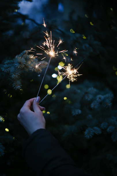 womans hand holding sparklers on the background of a Christmas tree with lights - Fotografie, Obrázek